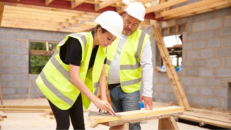 Kvinnelig bygningsarbeider tegner opp på en planke, mens mannlig bygningsarbeider overværer.