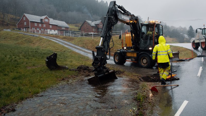 Traktor graver i en delvis oversvømt grøft, med et gårdsbruk i bakgrunnen. 