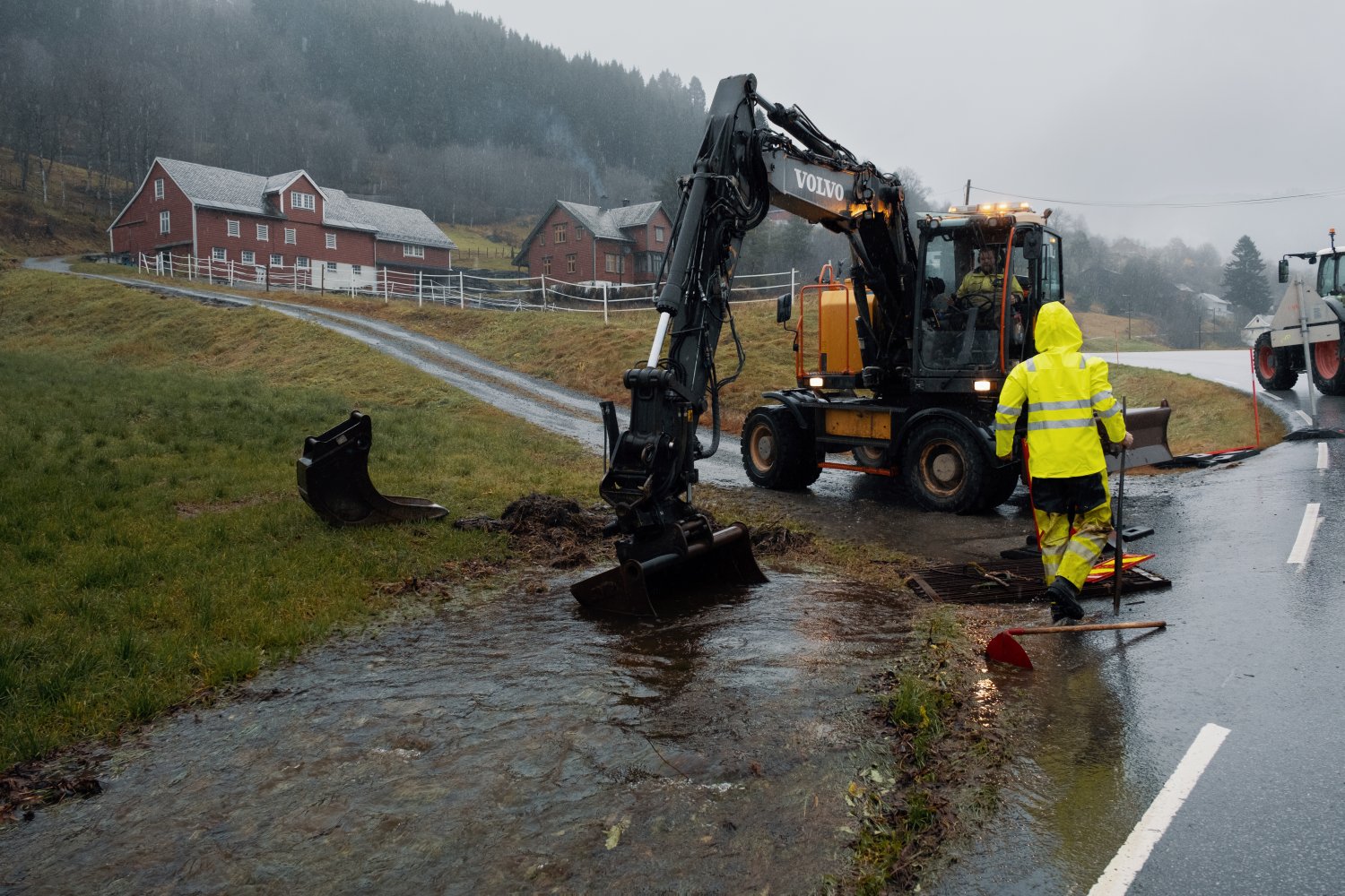 Traktor graver i en delvis oversvømt grøft, med et gårdsbruk i bakgrunnen. 